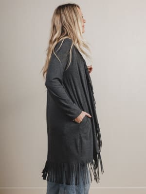 Woman wearing a long dark gray cardigan with fringe details on a plain background