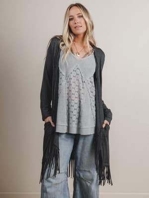 Woman wearing a gray cardigan with fringe, light blue top, and jeans against a plain background