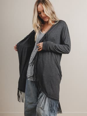 Woman wearing a gray cardigan with black fringe over a plaid shirt and jeans.