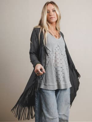 Woman wearing a long dark gray cardigan with fringe, layered over a light gray top and jeans, on a beige background.