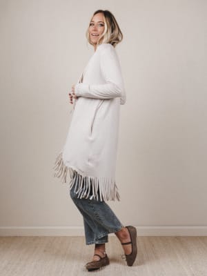 Woman wearing a long white cardigan with fringe, standing against a plain background
