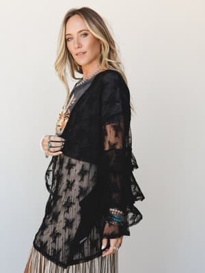 A sheer black lace kimono with dramatic ruffle bell sleeves layered over a floral graphic tee and pleated cream skirt — the kind of outfit that looks like it has a story.