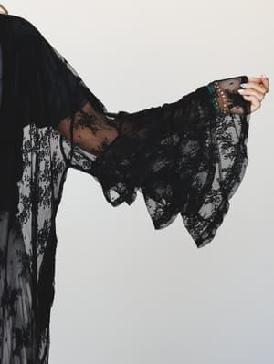 A close-up of a black floral lace kimono's dramatic bell sleeve, arm raised with stacked beaded bracelets peeking through the sheer, floaty fabric — the kind of piece that makes a regular outfit feel like a whole moment.