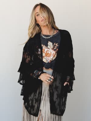 A layered boho look built around a black sheer lace kimono with tiered ruffle sleeves, worn open over a graphic floral tee and a cream fringe skirt — the whole thing accessorized with stacked silver rings, beaded bracelets, and a coin necklace for th