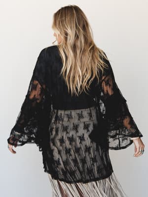 Shot from behind, a model wears a dramatic black floral lace kimono with sheer bell sleeves over what appears to be a cream fringe skirt — the whole look has that effortless dark-boho energy, like she's mid-twirl on her way somewhere interesting.