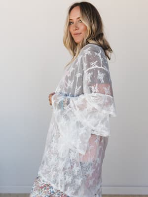 A sheer white floral lace kimono layered over a floral midi skirt, styled with stacked bracelets — the kind of piece that makes everything underneath feel intentional.