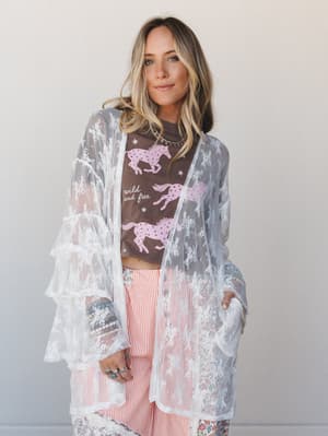 A sheer off-white floral lace kimono with tiered ruffle bell sleeves styled open over a brown horse graphic tee and red striped wide-leg pants — free-spirited layering with a nostalgic, expressive energy.