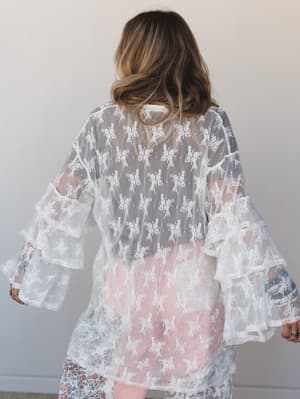 Shot from behind, a model wears a sheer off-white lace kimono with tiered ruffle bell sleeves over a red striped skirt and floral bottom — romantic layering with an effortless, almost bridal-boho energy.