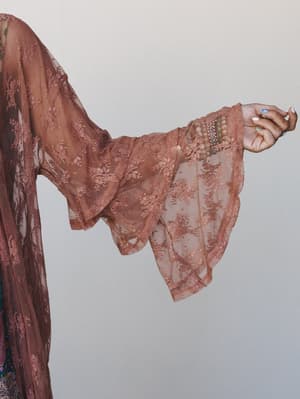 A close-up detail shot of a rose brown lace kimono's dramatic bell sleeve, catching light against a neutral studio backdrop — the floral lace overlay and stacked bracelets at the wrist do all the talking.