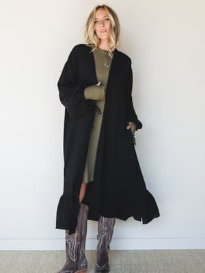A long black duster kimono layered over an olive ribbed midi dress, finished with tall western boots — the kind of outfit that looks effortless but is very much a decision.