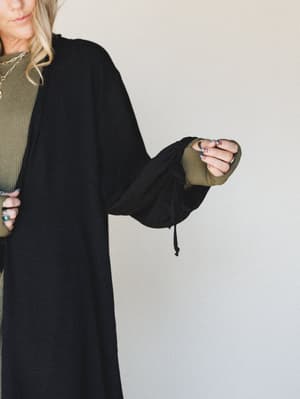 A close-up torso shot of a long black duster kimono layered over an olive ribbed top, with a drawstring detail at the sleeve hem — relaxed, effortless, and clearly meant for someone who layers on purpose.