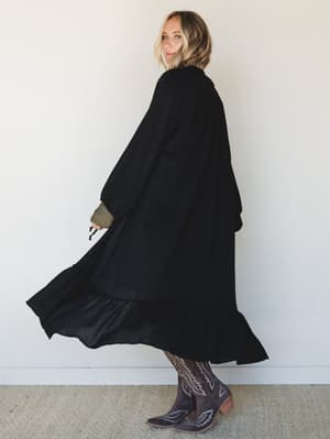 A flowing black duster kimono sweeps mid-twirl against a clean white wall, styled over an olive long-sleeve with embroidered western boots — the kind of outfit that feels equally at home at a farmers market or a late-night bonfire.