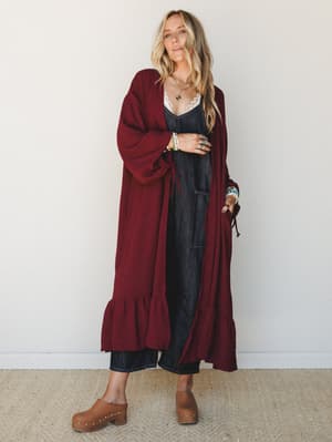A long, flowy wine-colored duster kimono with ruffle hem and tie cuffs layered over a dark denim jumpsuit and styled with wood-studded clogs and layered jewelry — the kind of outfit that makes a Tuesday feel intentional.