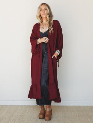 A long wine-red duster kimono with ruffle hem and cinched sleeves layered over a dark denim midi dress — the kind of outfit that feels intentional without trying too hard, styled with stacked bracelets and wood-platform clogs.