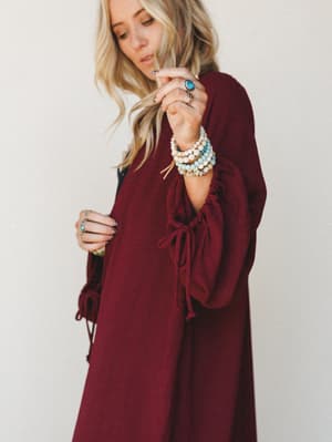 A wine-colored duster kimono with dramatic bell sleeves and tie cuffs, styled with stacked turquoise and cream beaded bracelets and silver rings — the kind of piece that feels intentional without trying too hard.