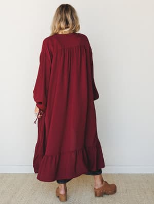 A back-facing model wears a voluminous wine-colored duster kimono with gathered shoulders and a ruffled hem, paired with dark jeans and tan wooden clogs — the kind of piece that makes a Tuesday feel intentional.