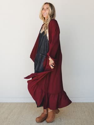 A smiling model shot in profile wears a floor-length wine-red ruffle-hem duster kimono layered over a dark denim jumpsuit with tan studded clogs — the kind of outfit that feels effortless and intentional at the same time.