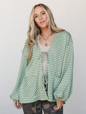 A relaxed gingham cardigan in avocado green with tie-front closures and a peplum hem, layered over a cream tank and camo pants — casual and a little unexpected in the best way.