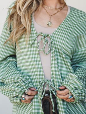 A green gingham cardigan with playful tie-front closures is layered over a white tank, styled with stacked silver rings and gold layered necklaces — the kind of outfit that looks effortless but was absolutely thought about.