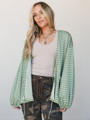 A relaxed, lived-in layering moment — a green gingham open cardigan worn loose over a blush tank and camo cargo pants, styled with layered gold necklaces for that effortless 'threw it on and it just worked' energy.