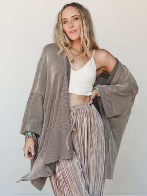 A relaxed, earthy outfit layered with intention — an oversized striped duster cardigan worn open over a white crochet bralette and pleated wide-leg pants, accessorized with turquoise stack bracelets and layered gold necklaces for that effortless boho