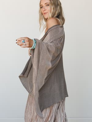 A model shot in three-quarter profile wearing a flowy open-front duster cardigan in a camel and black fine-stripe knit, styled over a white tank with wide-leg bottoms, accessorized with turquoise jewelry for an effortlessly relaxed, boho feel.