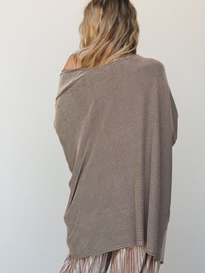 Shot from behind, a relaxed oversized knit top in a warm camel and dark stripe texture sits off one shoulder, layered over a pleated skirt — the whole look feels like a cozy Saturday that accidentally turned into something you'd actually wear out.