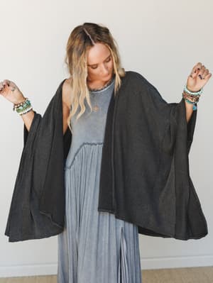 A flowy charcoal ribbed duster cardigan worn open over a washed blue maxi dress, styled with layered bracelets and a delicate pendant necklace — the kind of effortless layering that looks intentional without trying.