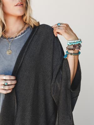 A close-up lifestyle shot of a ribbed charcoal duster cardigan layered over a faded blue tee, styled with a full stack of turquoise and stone bracelets, layered gold necklaces, and silver boho rings — the kind of effortless accessorizing that looks c