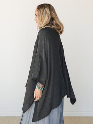 A draped, oversized duster cardigan in charcoal ribbed knit worn from a side angle over a dusty blue maxi skirt — the kind of layering piece that makes every outfit feel more intentional without trying too hard.