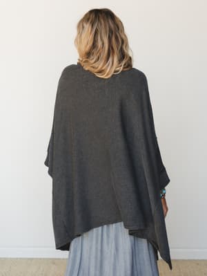 Shot from behind, a model wears an oversized knit poncho wrap in charcoal with fine stripe texture, layered over a flowy maxi skirt — the kind of effortless, cozy outfit that looks like it happened on purpose.