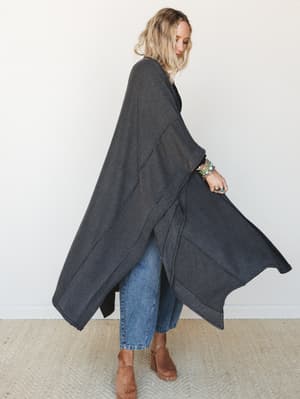 A model shot in profile wearing a dramatic floor-length ribbed duster kimono in charcoal over a black top and cropped jeans, styled with stacked turquoise bracelets and tan heeled clogs — the kind of layering piece that makes an ordinary Tuesday feel