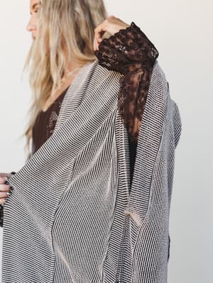 A close-up detail shot of a striped knit duster kimono with dramatic open sleeves, layered over a dark floral lace top — the contrast between the relaxed woven texture and the delicate lace underneath is doing a lot of quiet, interesting work.