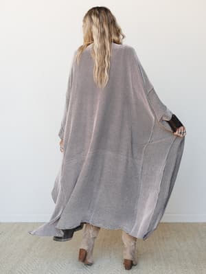 Shot from behind, a model in a floor-length open duster kimono with a semi-sheer striped knit texture lets the draped sleeves fan out, styled with tan cowboy boots and stacked turquoise rings — effortlessly boho and a little dramatic in the best way.