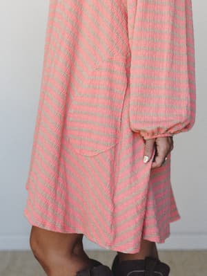 A close-up detail shot of a textured diagonal stripe dress in coral peach and warm tan, with a relaxed bishop sleeve cuffed at the wrist and a patch pocket — easy and a little retro in the best way.