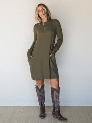A relaxed ribbed knit mini dress in dusty olive, styled with tall brown cowboy boots and a layered gold necklace — the kind of effortless outfit that works just as hard on a weekend errand run as it does at a fall bonfire.