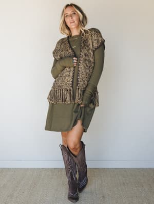 An olive ribbed sweater dress layered under a chunky fringe knit vest, finished with dark brown cowboy boots — the kind of fall outfit that looks effortless but took exactly the right instincts to pull together.