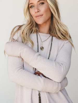 A soft, close-up lifestyle shot of a model in a ribbed long sleeve dress the color of warm cream, arms crossed loosely, layered necklaces catching the light — the kind of outfit that looks equally at home on a slow Sunday or a casual lunch out.