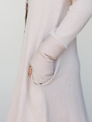 A close-up detail shot of a ribbed knit dress in a soft oat tone, hand tucked into a side pocket — the kind of cozy, lived-in piece you reach for without thinking twice.