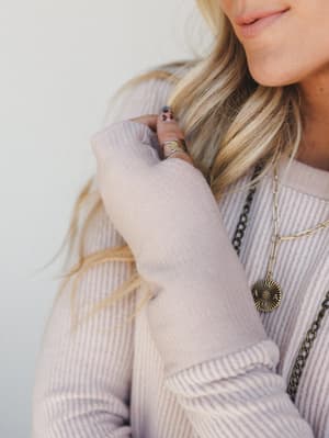A close-up lifestyle shot of a ribbed knit sweater dress in a soft blush-oat tone, styled with layered gold and antique brass necklaces and a silver ring — the kind of effortless, jewelry-heavy look that feels personal rather than put-together.