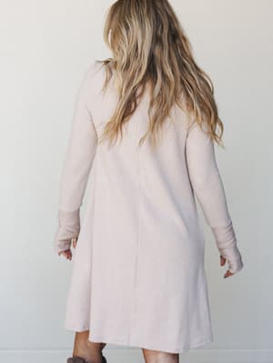 A soft ribbed knit mini dress in a barely-there blush oat tone, shot from behind — the relaxed A-line silhouette and thumbhole cuffs give it that cozy, lived-in feel that's just as happy on a coffee run as it is on the couch.