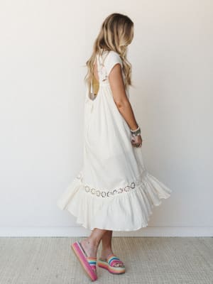 A breezy cream midi dress shot from behind, showing off an open back with crochet trim detail and a ruffled hem — styled with rainbow platform sandals and stacked boho bracelets for that effortless summer energy.
