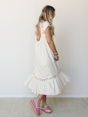A breezy cream midi dress shot from behind, showing off an open back with crochet trim detail and a ruffled hem — styled with rainbow platform sandals and stacked boho bracelets for that effortless summer energy.