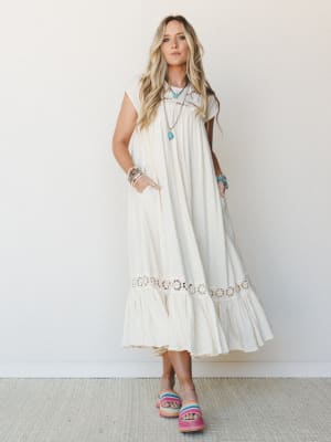 A flowy cream midi dress with crochet trim details and a ruffled hem, styled with turquoise jewelry and colorful woven platform sandals against a soft studio wall — effortlessly boho and made for warm-weather wandering.