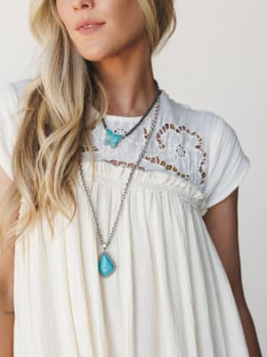 A cream gauze dress with floral crochet yoke detailing, styled with layered turquoise stone necklaces — the kind of effortless boho look that feels equally at home on a porch or a summer road trip.