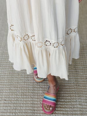 A close-up of a cream gauze maxi dress hem with delicate daisy lace trim and a ruffled bottom tier, paired with colorful rainbow-striped woven sandals — the kind of detail shot that makes you want to reach out and touch it.