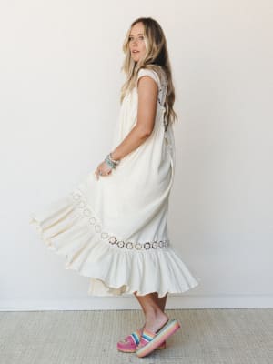A cream midi dress twirls mid-motion against a soft white studio wall — the floral crochet hem trim and ruffle skirt doing exactly what they're meant to, while rainbow platform slides and stacked turquoise bracelets keep it grounded and personal.