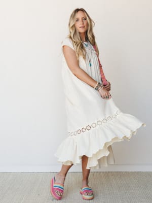 A flowy cream midi dress with crochet floral trim and a ruffled hem catches mid-twirl, styled with rainbow platform sandals, layered turquoise jewelry, and a colorful woven crossbody — the kind of outfit that feels like a good mood.