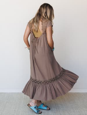 Shot from the back, a flowy mocha maxi dress fans out mid-twirl, showing off its open lace back, crochet hem band, and ruffled skirt — styled with a mustard bralette peeking through and turquoise sandals for that effortlessly layered, lived-in boho f