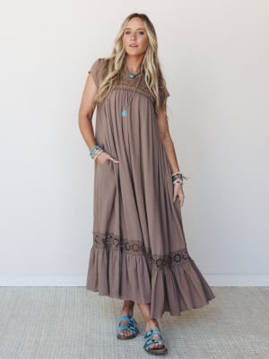 A flowy mocha maxi dress with lace yoke and tiered ruffle hem, styled with turquoise sandals and stacked boho jewelry against a clean studio wall — the kind of dress that makes a farmers market feel like an occasion.